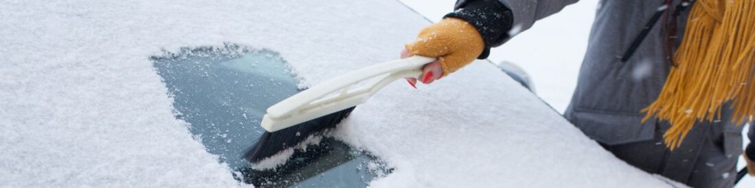 A person using an ice scraper to clear snow from a car windshield, emphasizing the necessity of winter travel essentials.